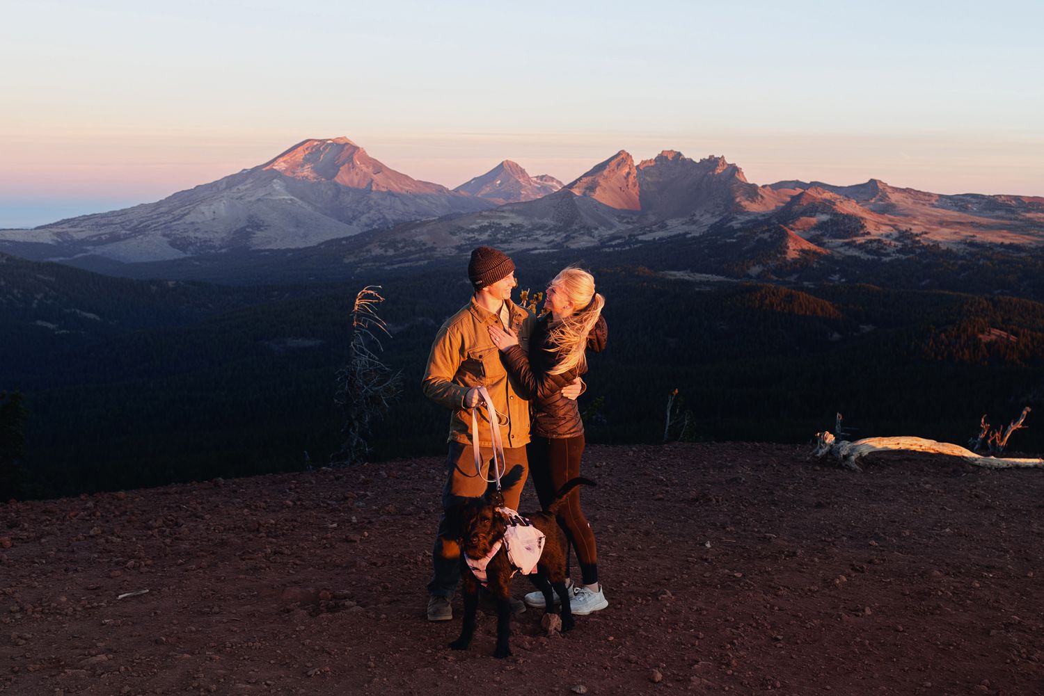 Couple embracing at a mountain summit at golden hour with a dog beside them and snow-capped peaks glowing in the background.