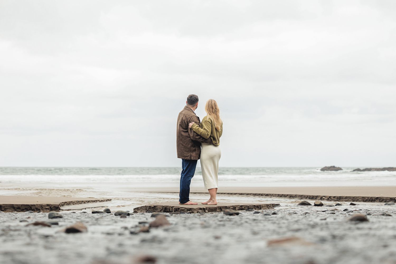 Couple standing together on a rocky beach gazing at the ocean on an overcast day.