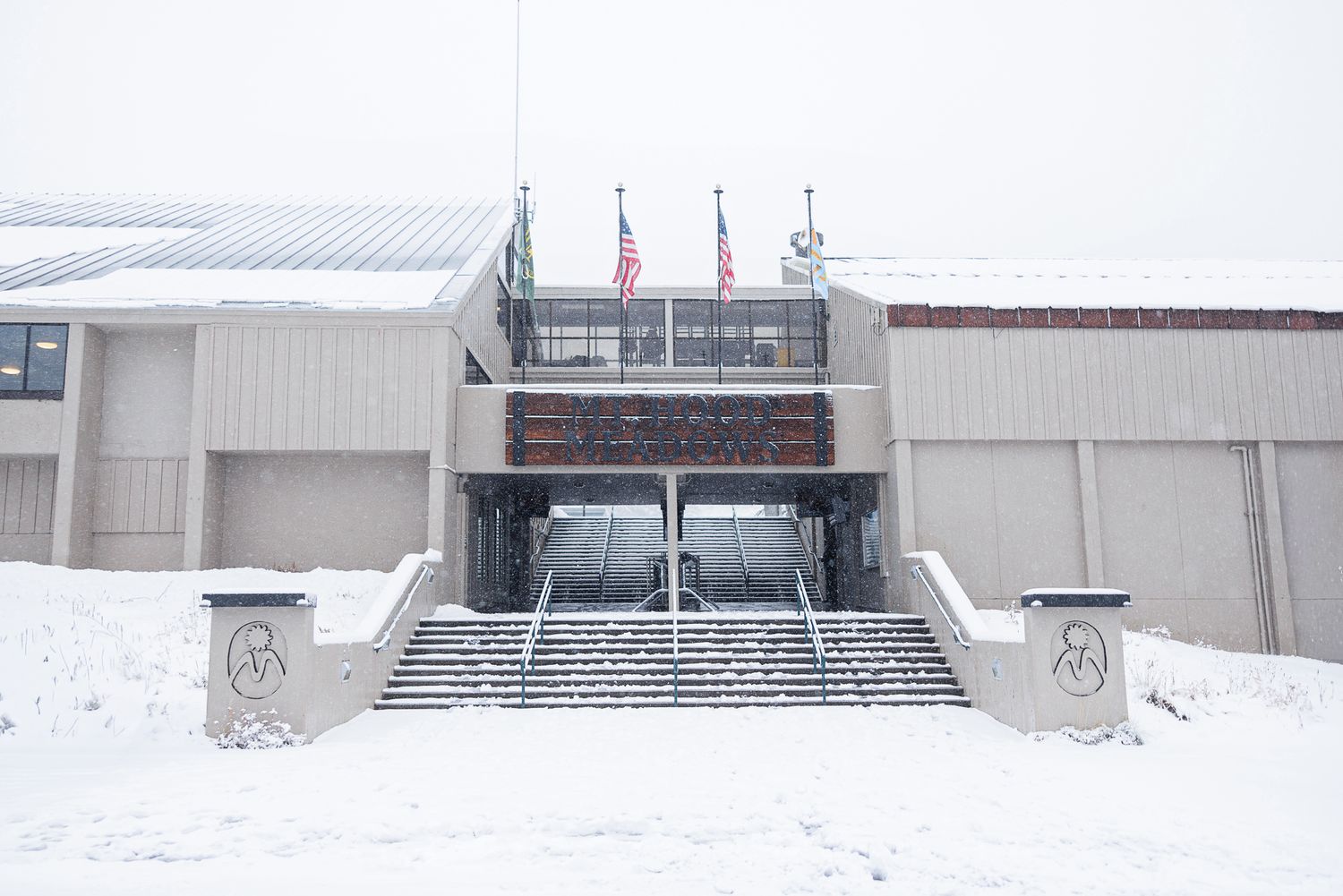 Snow-covered entrance stairs leading to a modern building with flags on top during winter weather.