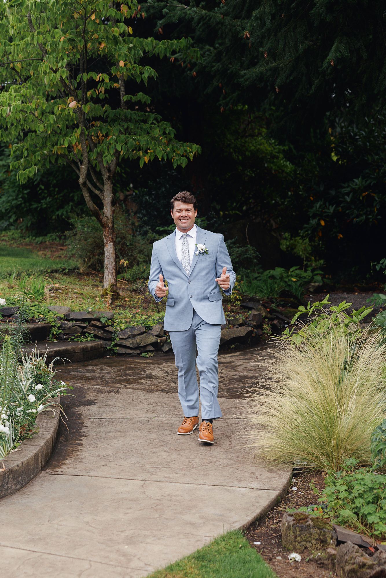 A person in a light blue suit walks along a curved garden path surrounded by ornamental grasses and trees.