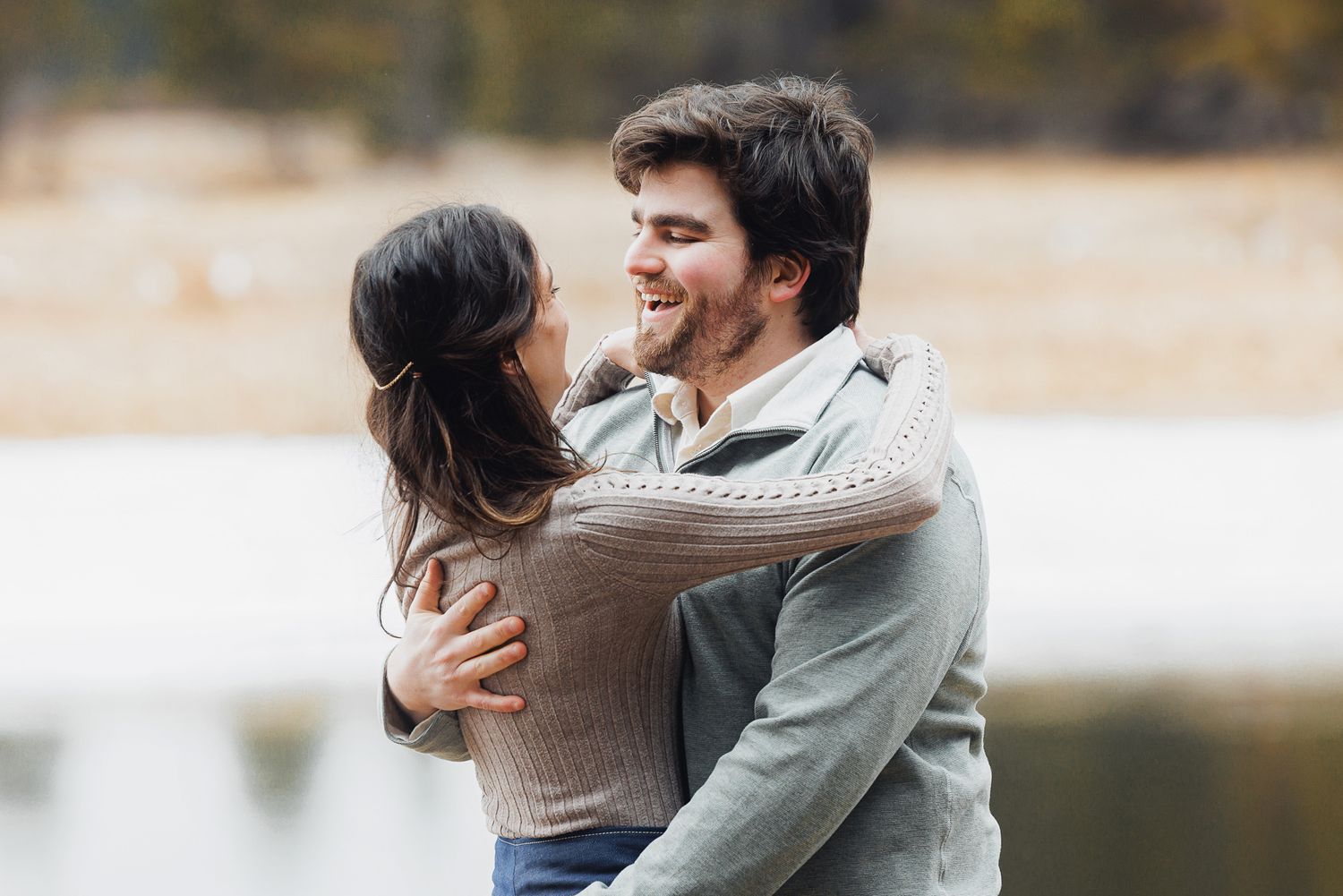 A couple embraces joyfully by a serene lake on an autumn day.