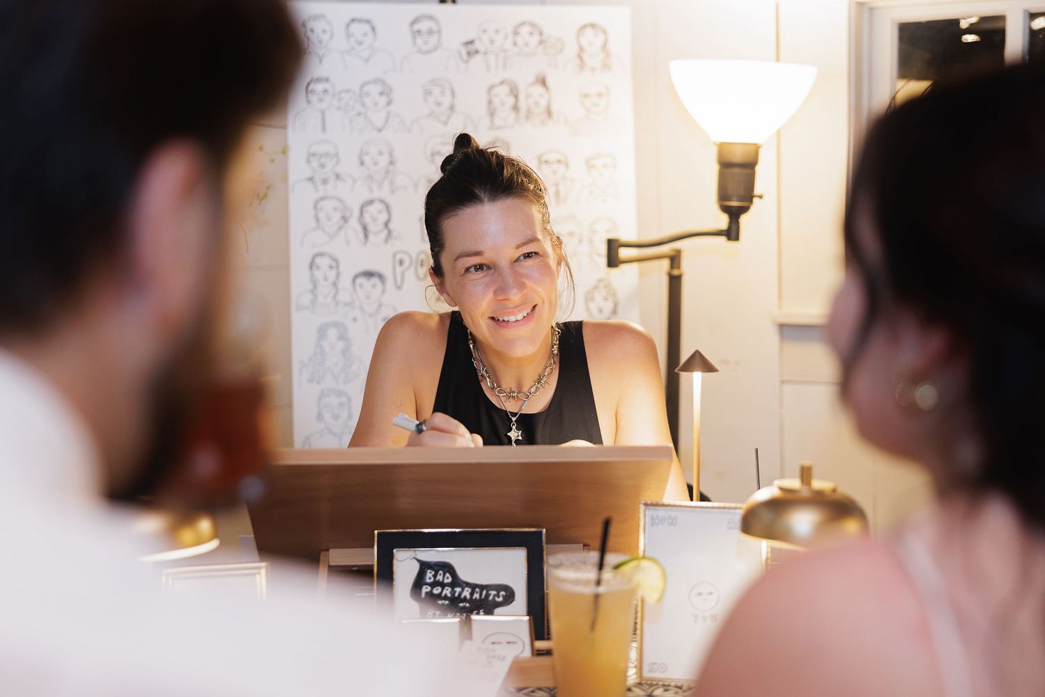 Smiling woman leaning on counter with laptop in cozy cafe, chatting with customers near warm lamp light.