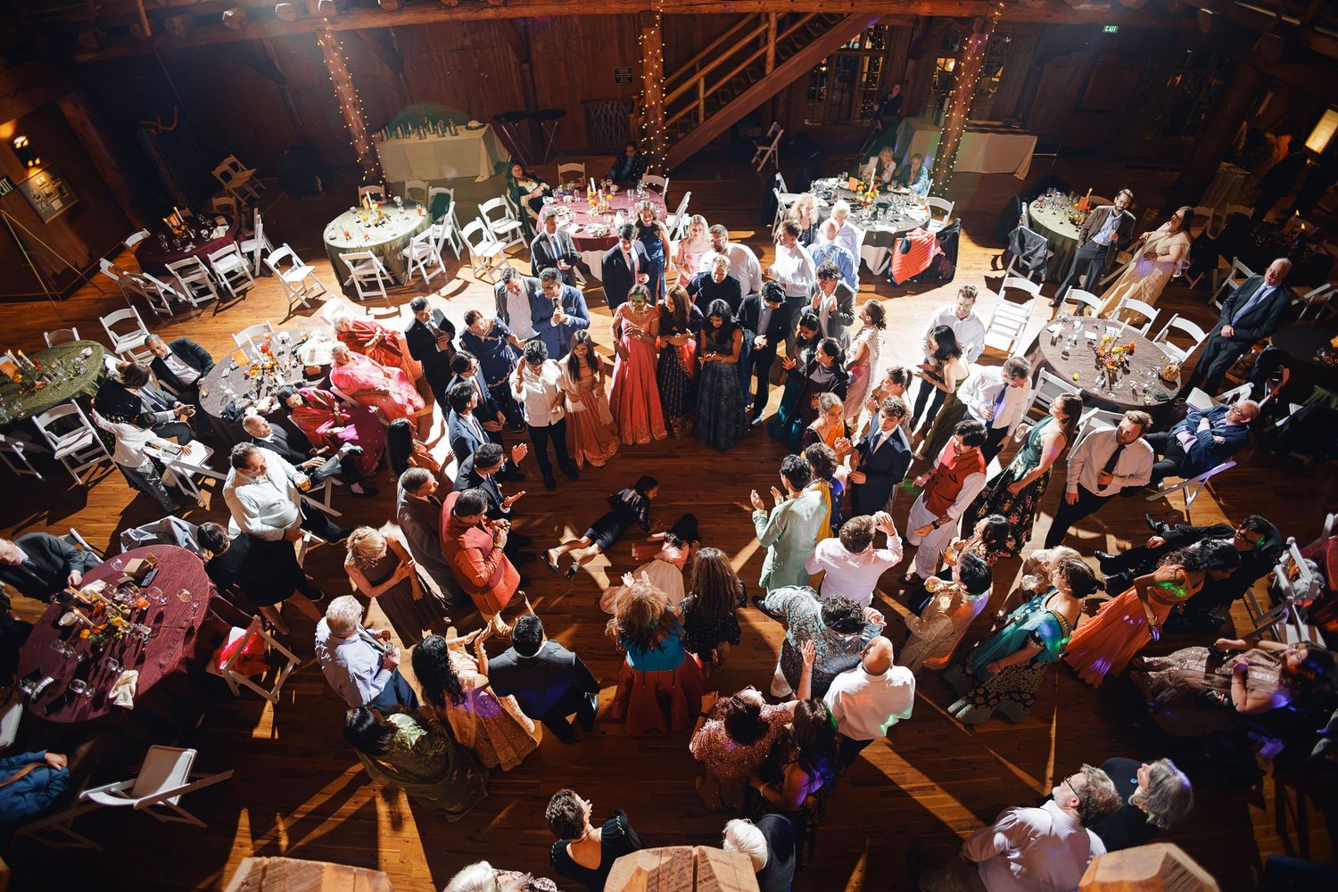 Aerial view of a lively wedding reception with guests dancing and mingling on a wooden dance floor indoors.