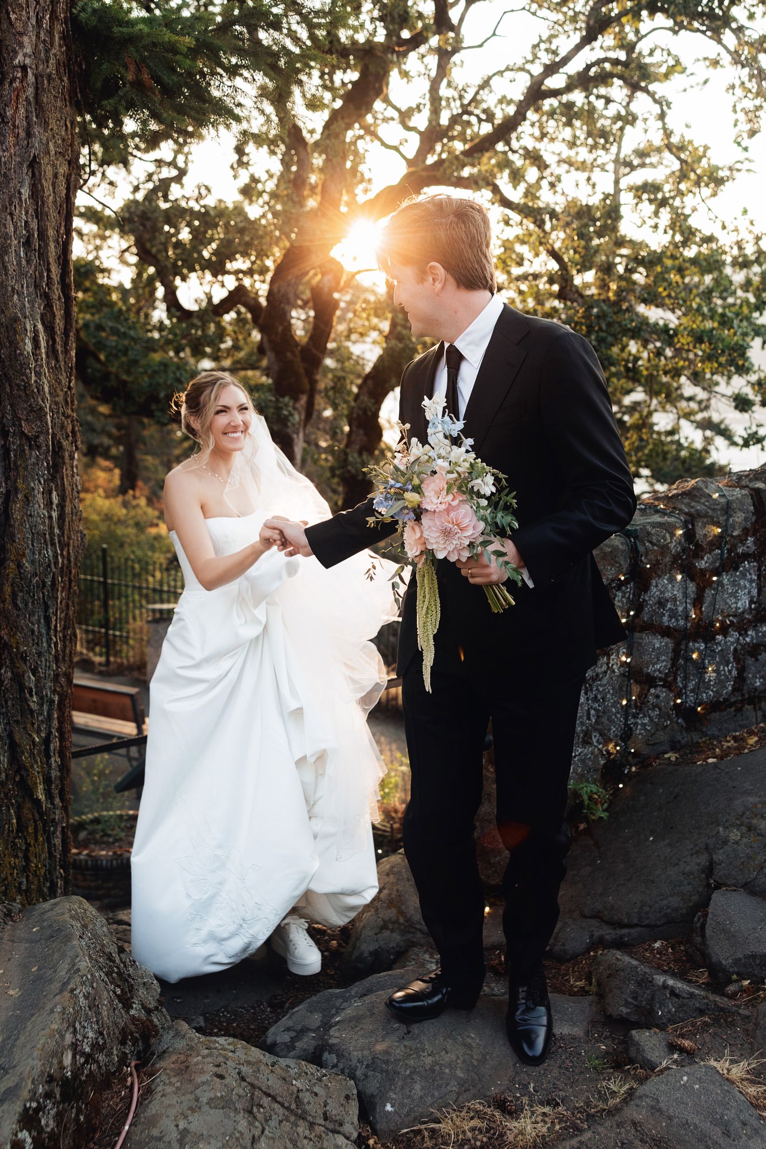 Bride and groom share a joyful moment outdoors at sunset, with bouquet in hand, surrounded by trees and rocks.