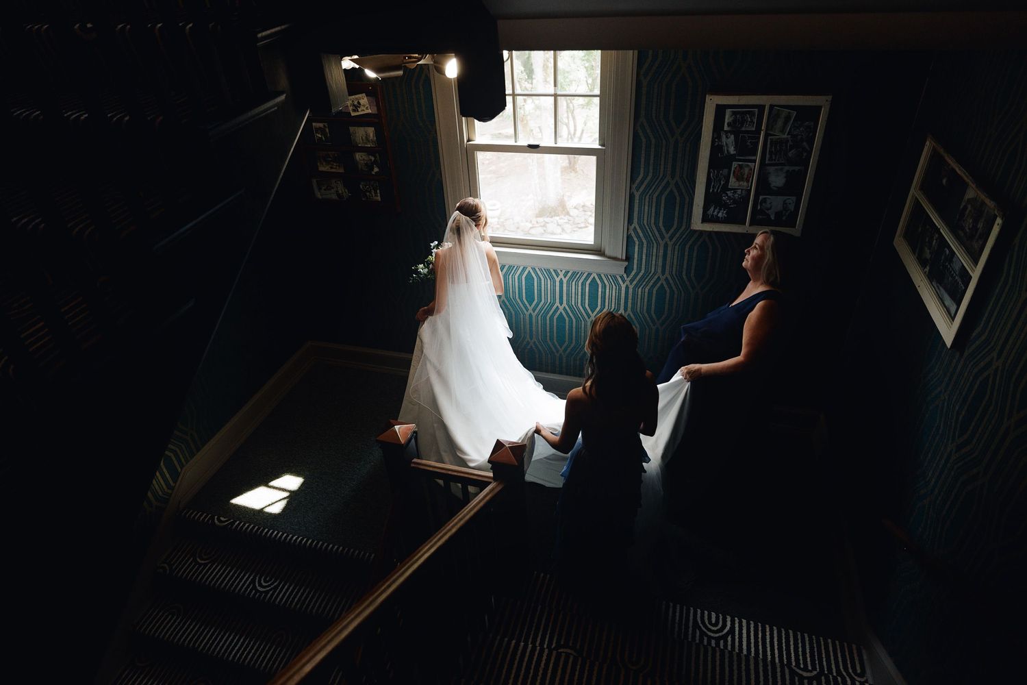 Bride in white gown stands by window on staircase, assisted by bridesmaids in dark elegant venue.