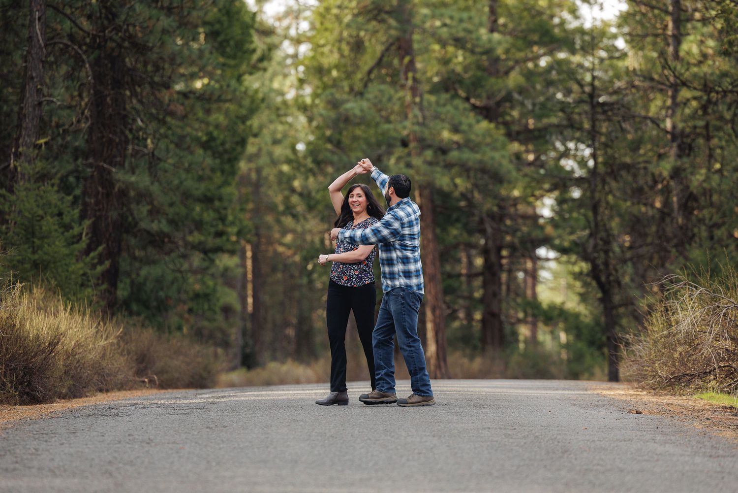 Couple dancing together on a tree-lined road in a forest, woman twirling under mans raised arm.