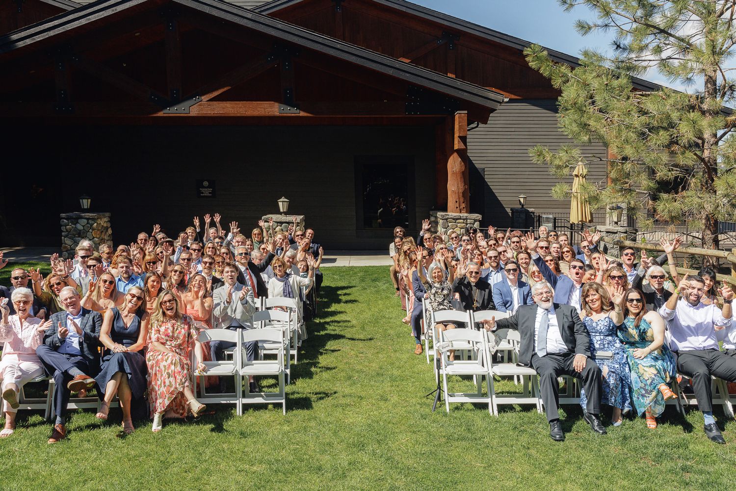 Large group of wedding guests seated in white chairs on green lawn outside a rustic wooden venue on a sunny day.