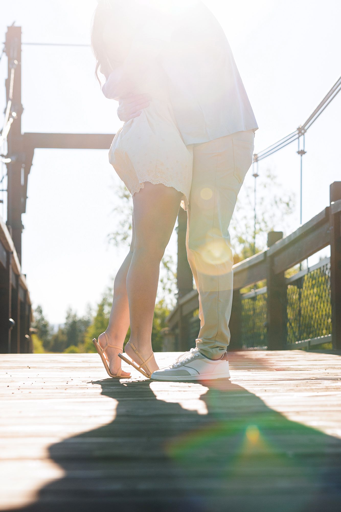 Couple standing close together on a wooden bridge in bright sunlight, wearing casual white and khaki outfits.