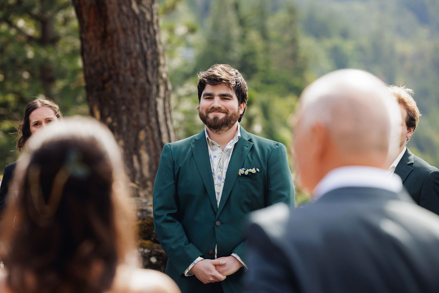 Groom in green suit smiling at outdoor wedding ceremony surrounded by guests and pine trees.