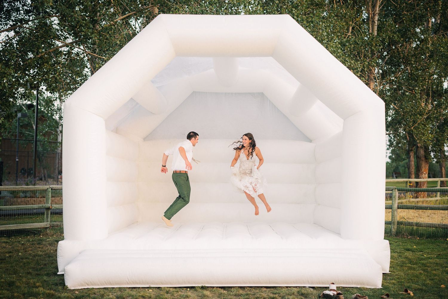 A couple jumps playfully inside a white inflatable bounce house at an outdoor wedding venue.