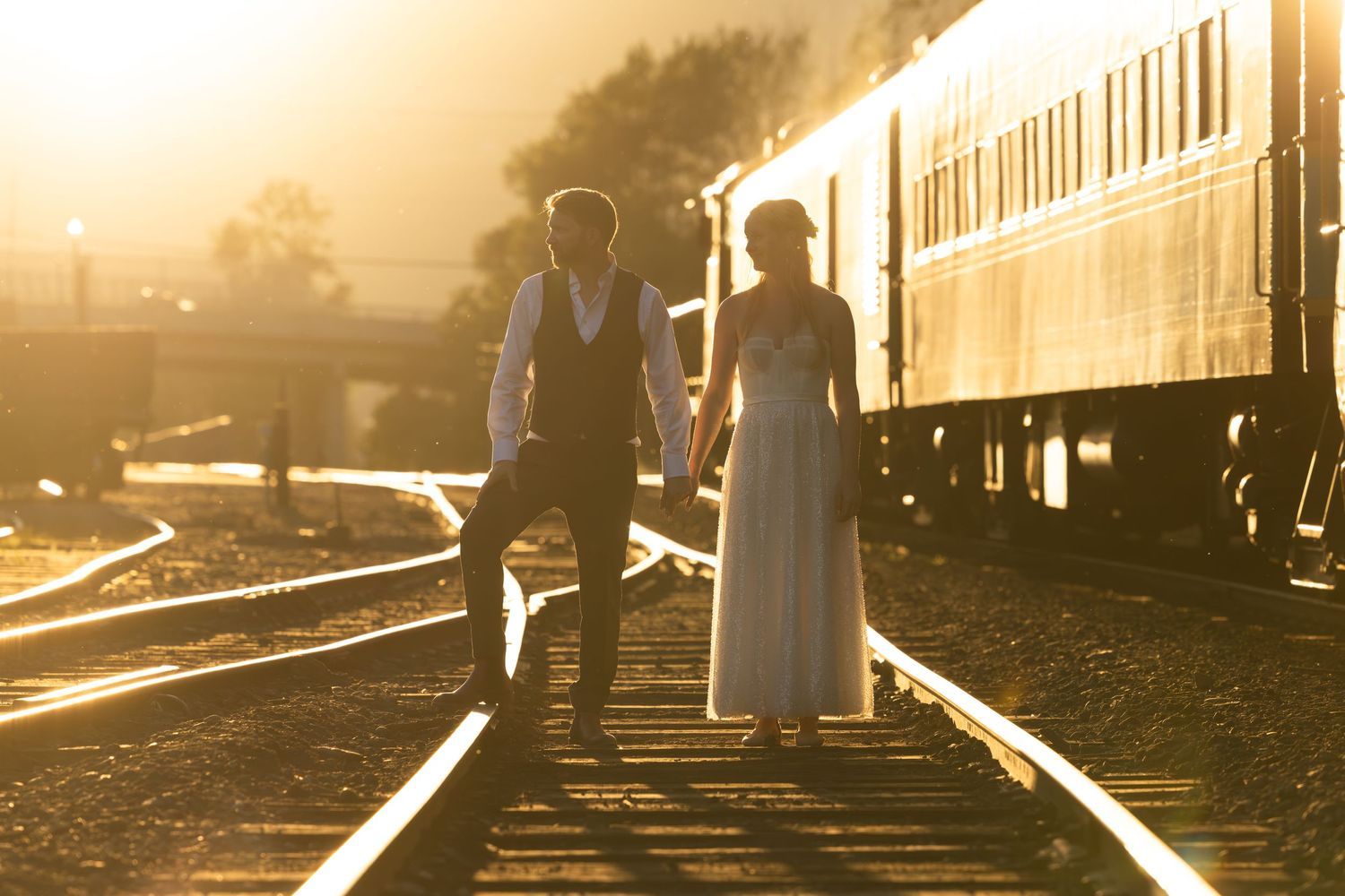 A couple in wedding attire stands on train tracks at sunset with a vintage railcar in the background.
