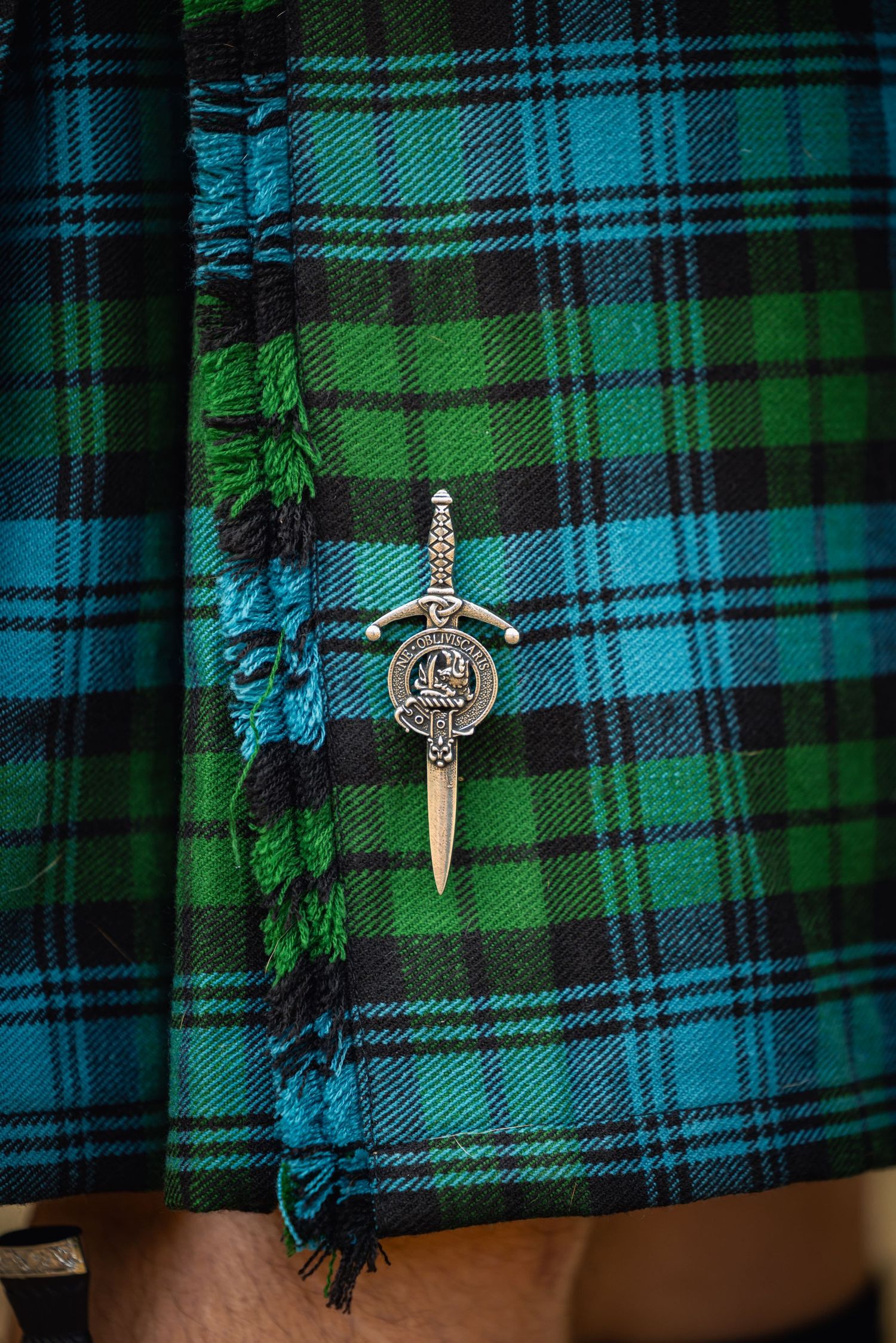 Traditional Scottish kilt pin with Celtic design against green and blue tartan plaid fabric with fringe detail.