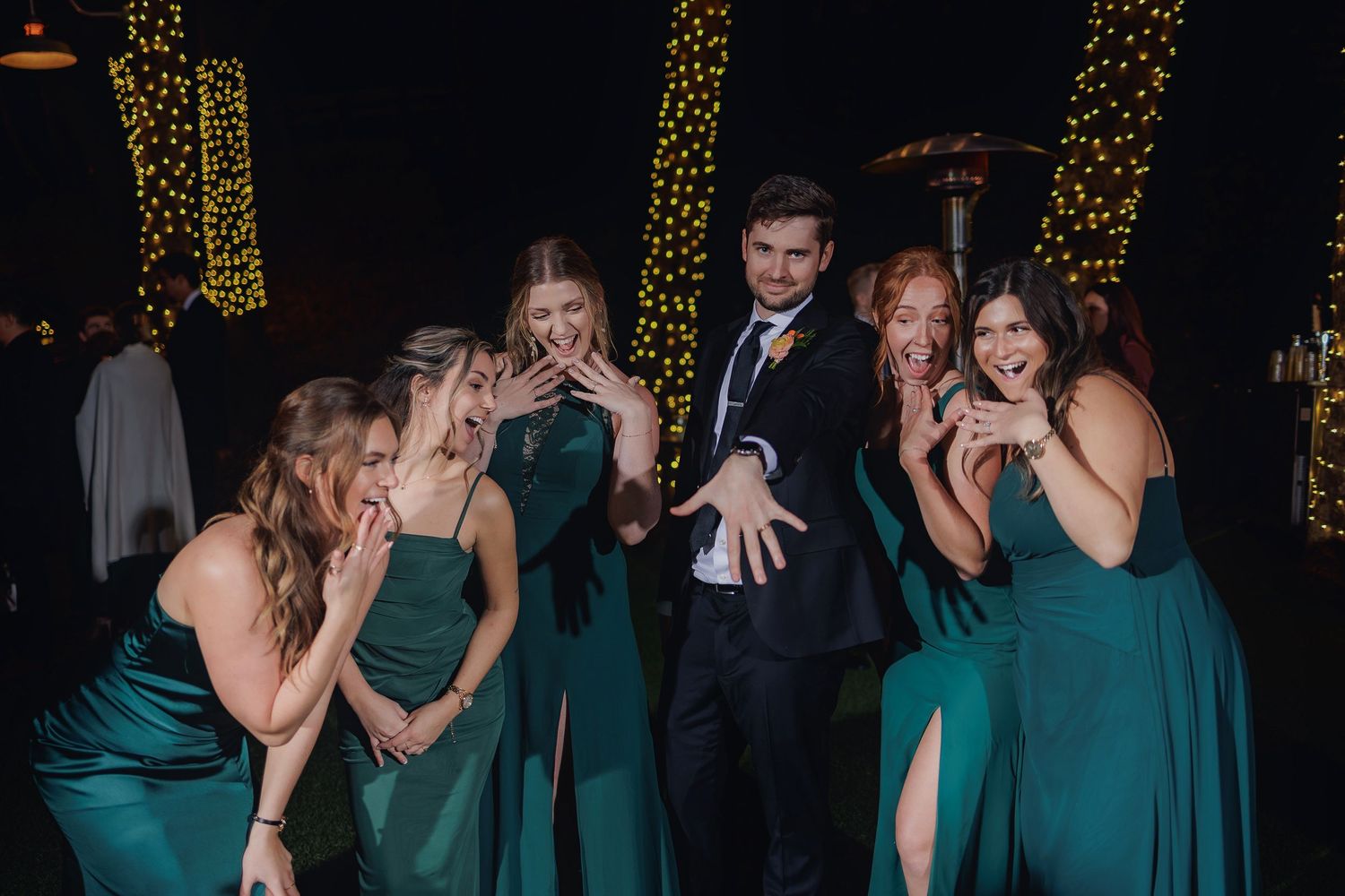 Wedding party in formal attire poses together with expressions of surprise against a backdrop of twinkling lights.