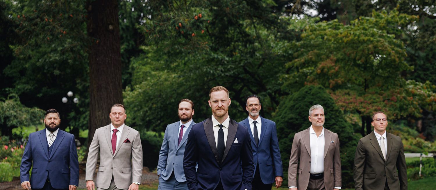 Group of seven well-dressed men in suits posing outdoors in a lush green garden setting for a formal occasion.