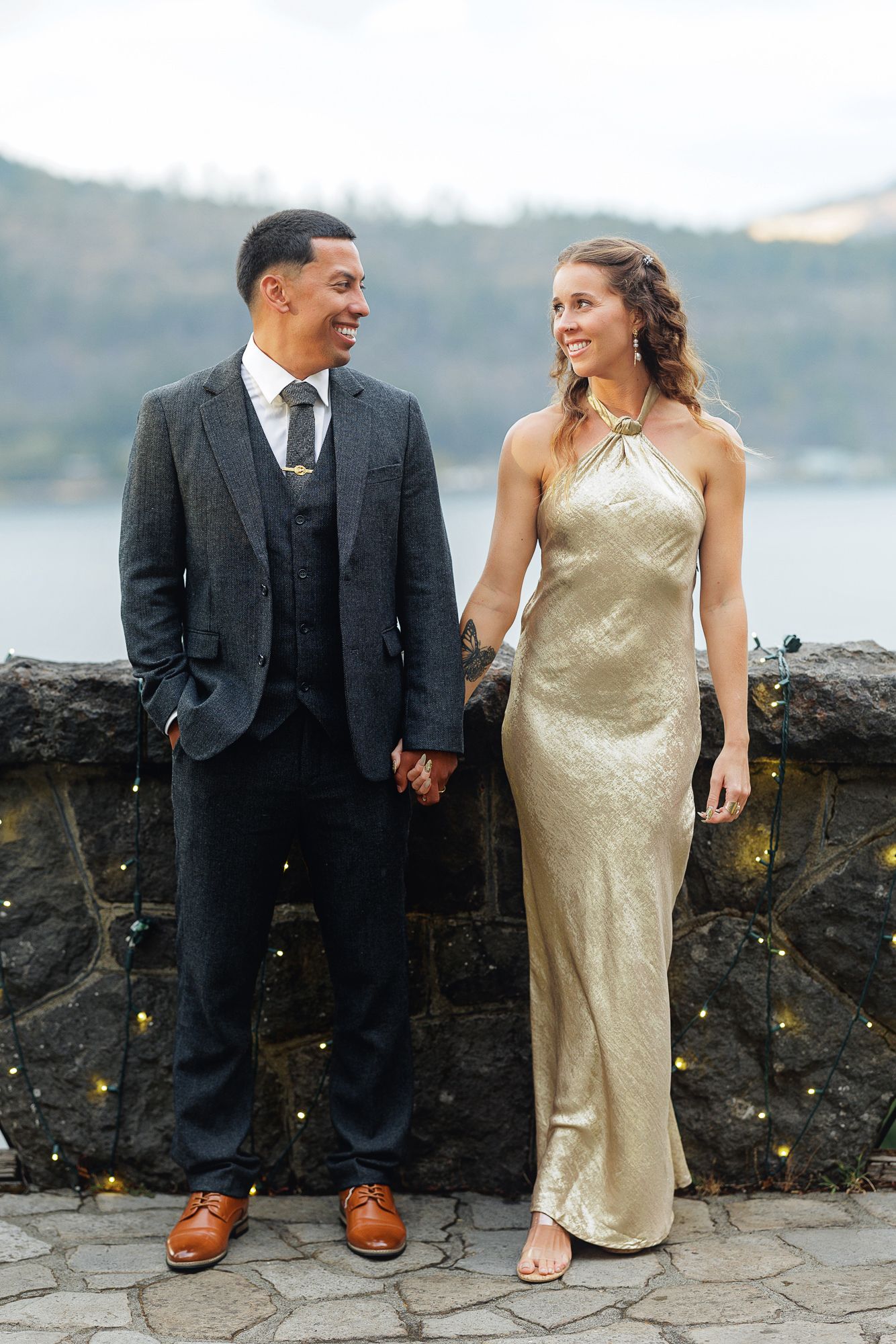 Elegant couple holding hands outdoors, man in gray suit, woman in gold halter gown, with scenic water view backdrop.