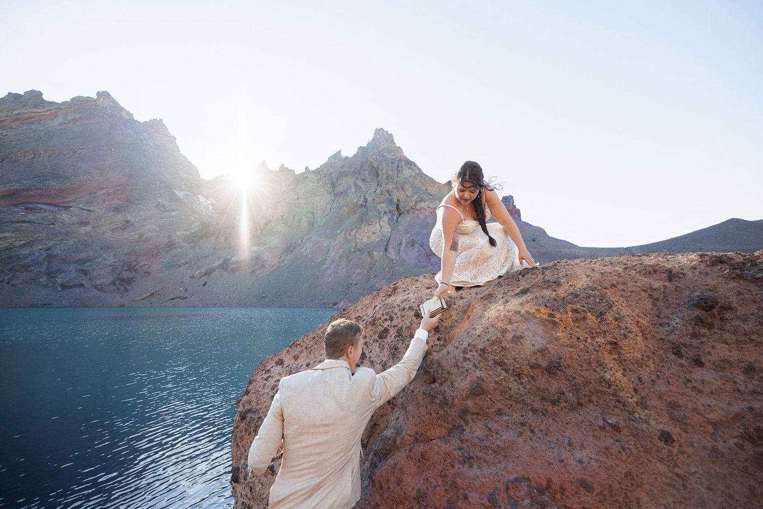 Bride in a white gown and groom in a cream suit hold hands while climbing rugged red rocks beside a blue lake at sunset.