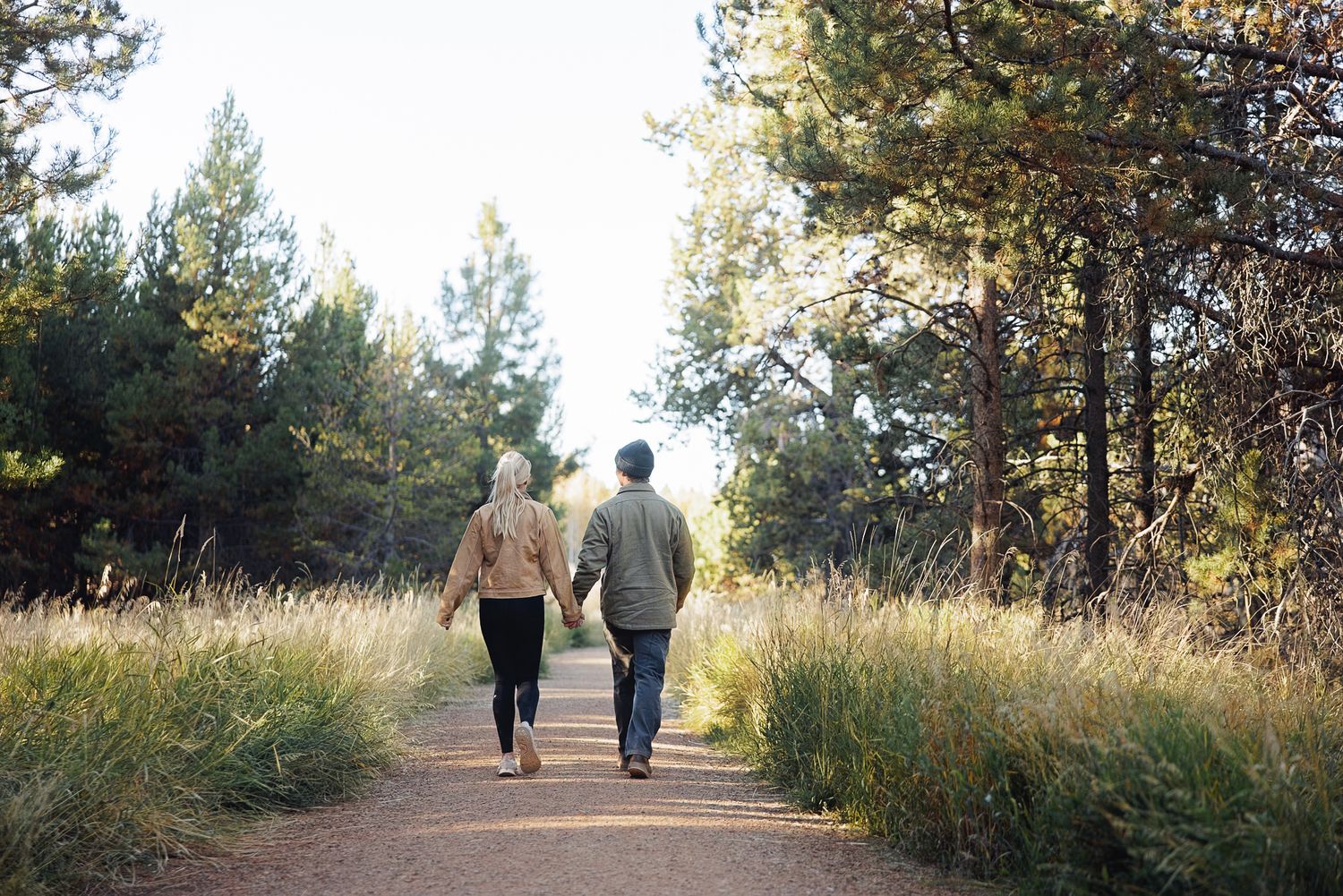 Couple holding hands while walking along a dirt trail through tall golden grass and pine trees on a peaceful autumn day.