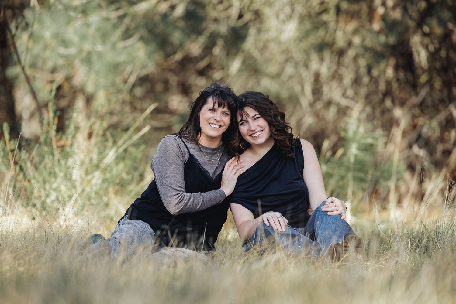 Two women sitting together in a grassy field, smiling warmly, with trees in the background.