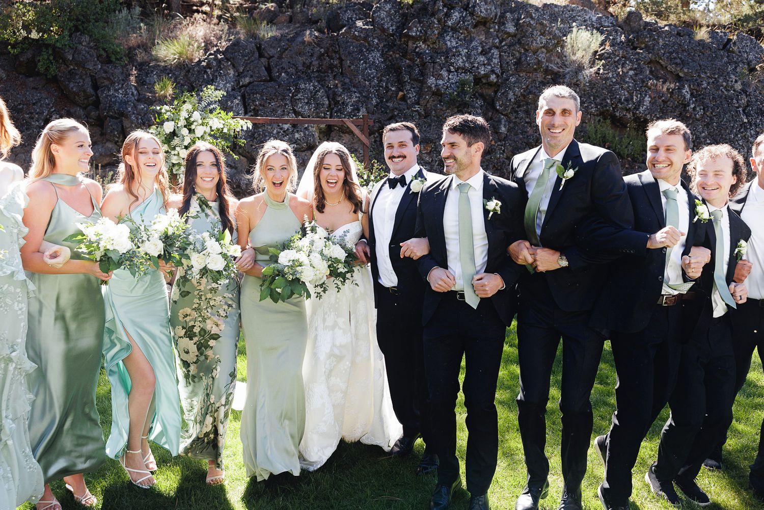 Wedding party in black tuxedos and sage green bridesmaid dresses pose together outdoors, laughing joyfully.