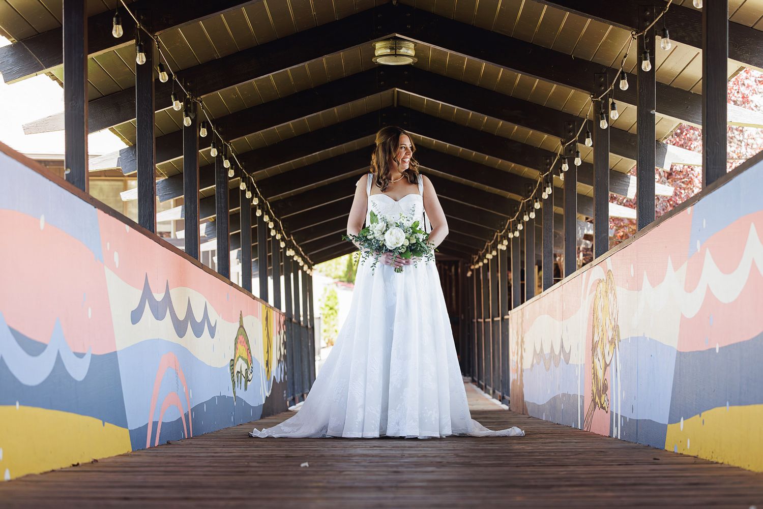 Bride in white gown holds bouquet on colorful mural-adorned covered wooden bridge, elegant wedding portrait.