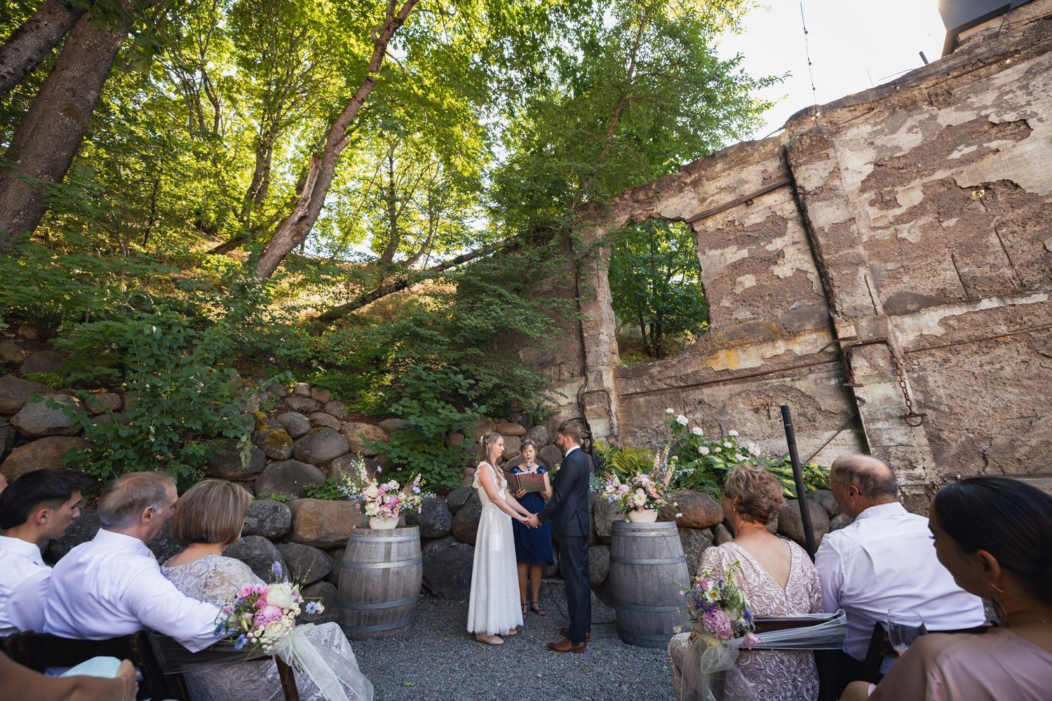 Intimate outdoor wedding ceremony amongst stone walls and trees with guests seated on benches.