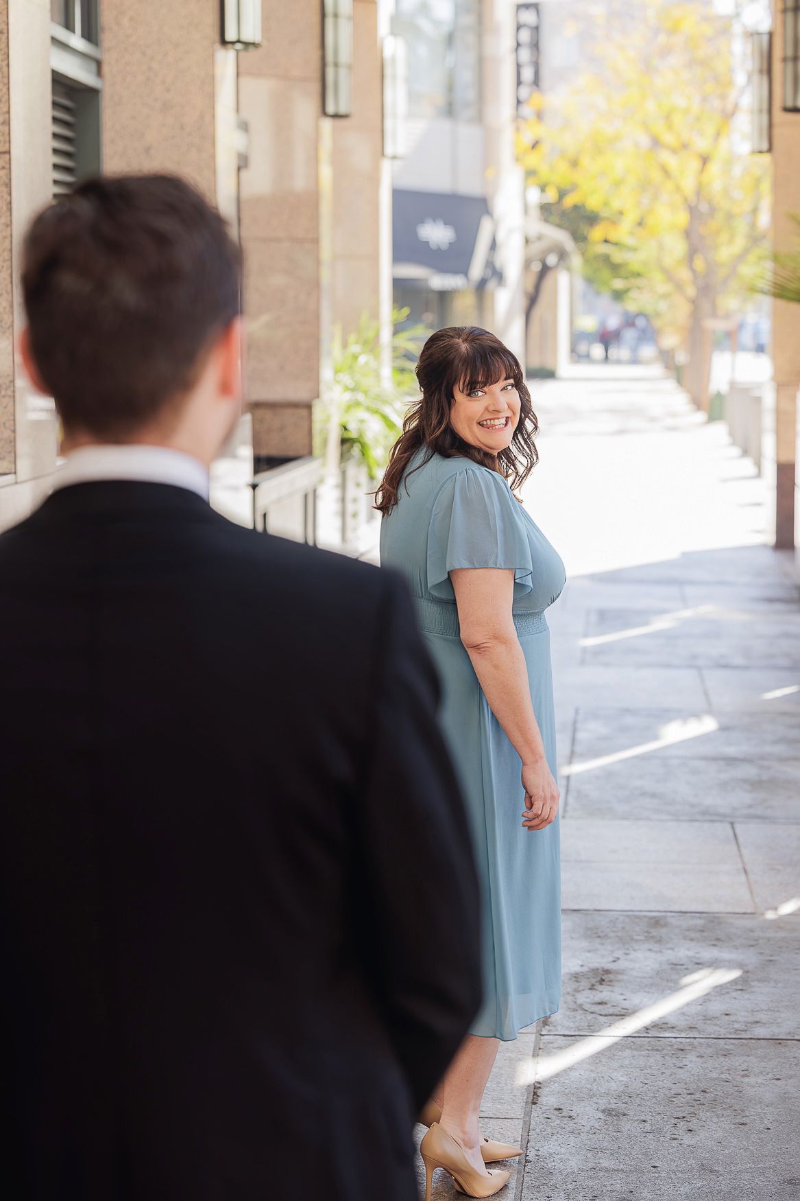 A couple stands in an urban setting with sunlit trees in the background as a romantic moment unfolds.