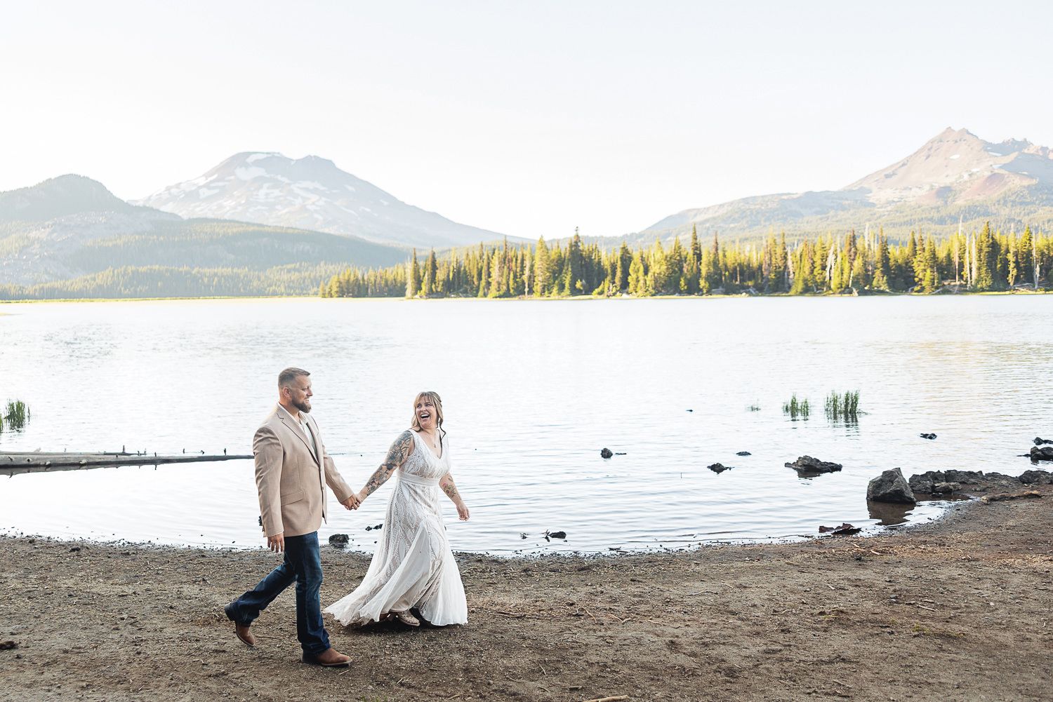 A couple walks along a scenic mountain lake beach during sunset with tall evergreen trees in the background.