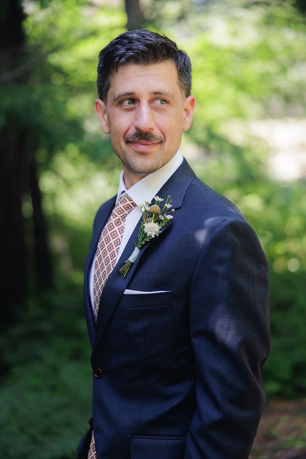 A wedding attendee in a navy suit and patterned tie stands in a forest setting, wearing a boutonniere on their lapel.