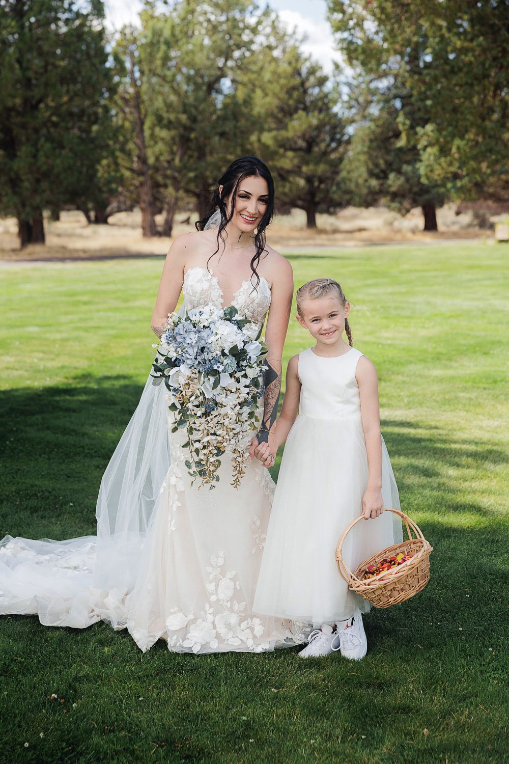 A wedding dress and delicate white flower girl dress stand out against lush green grass in an outdoor garden setting.