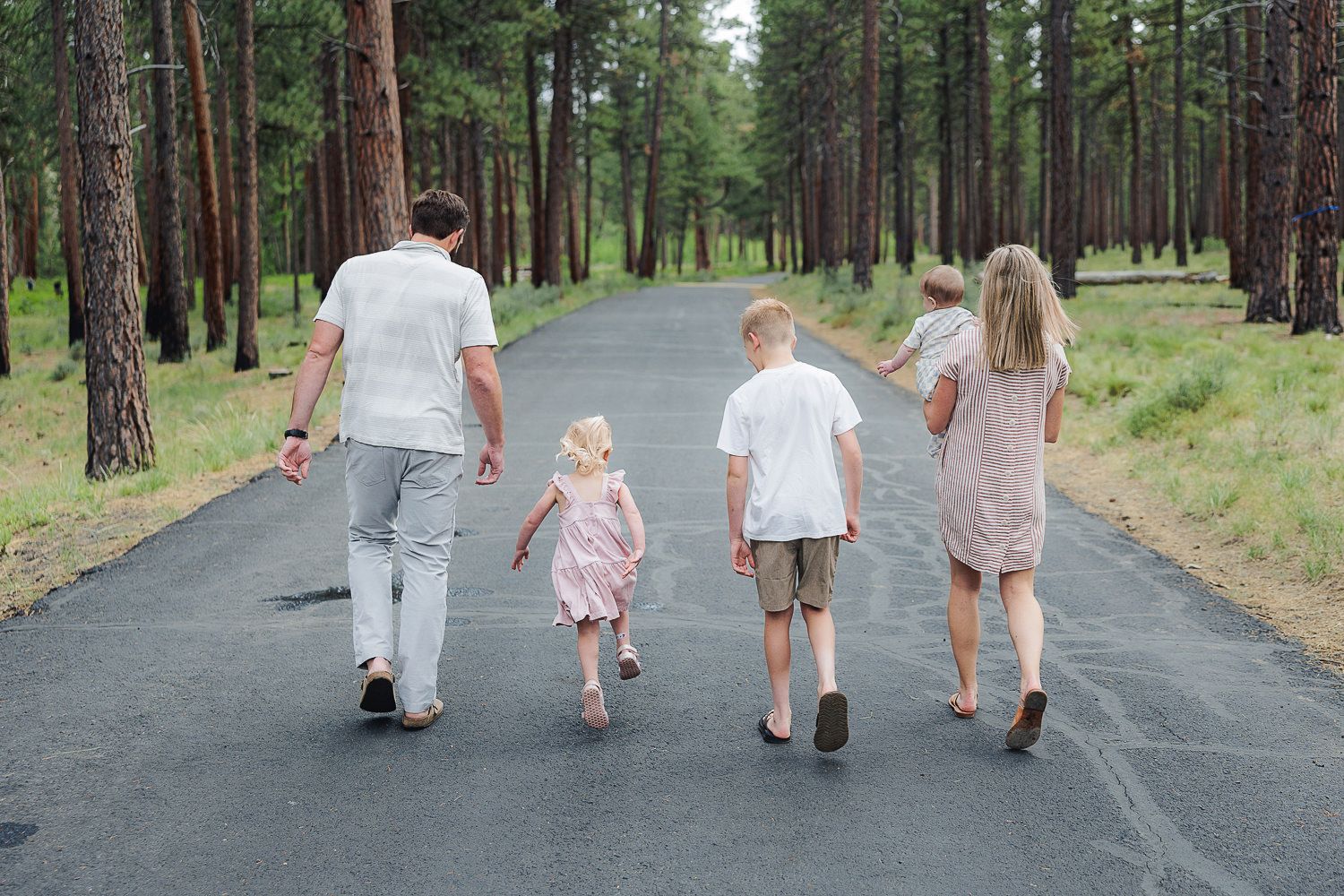 Family members hold hands while walking together down a tree-lined paved road in a peaceful forest setting.