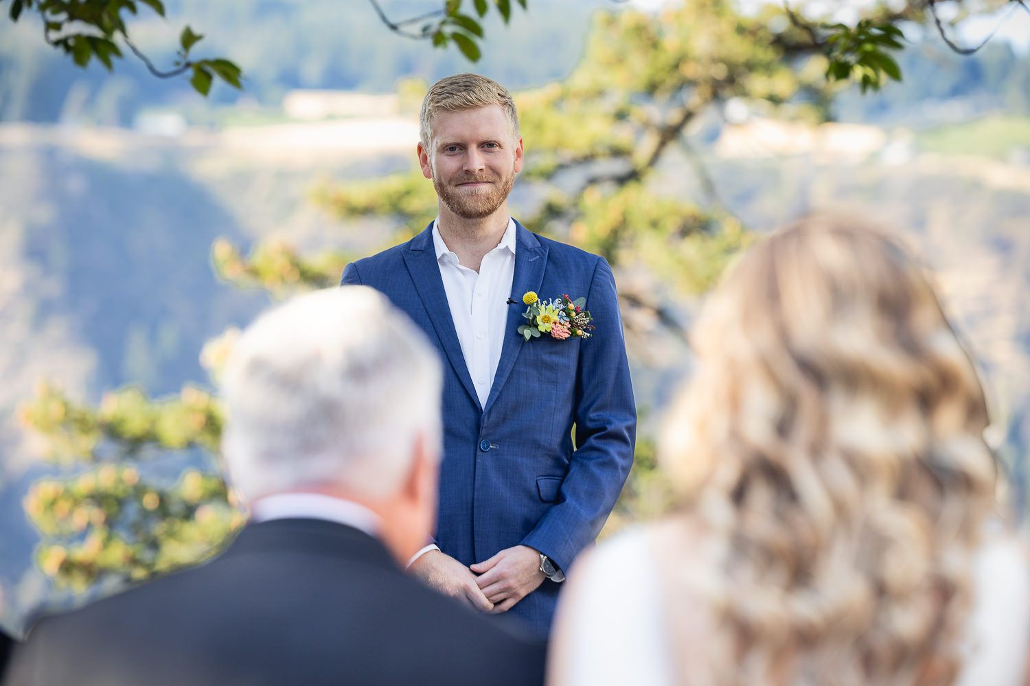 A groom in a navy suit with boutonniere smiles during an outdoor wedding ceremony.