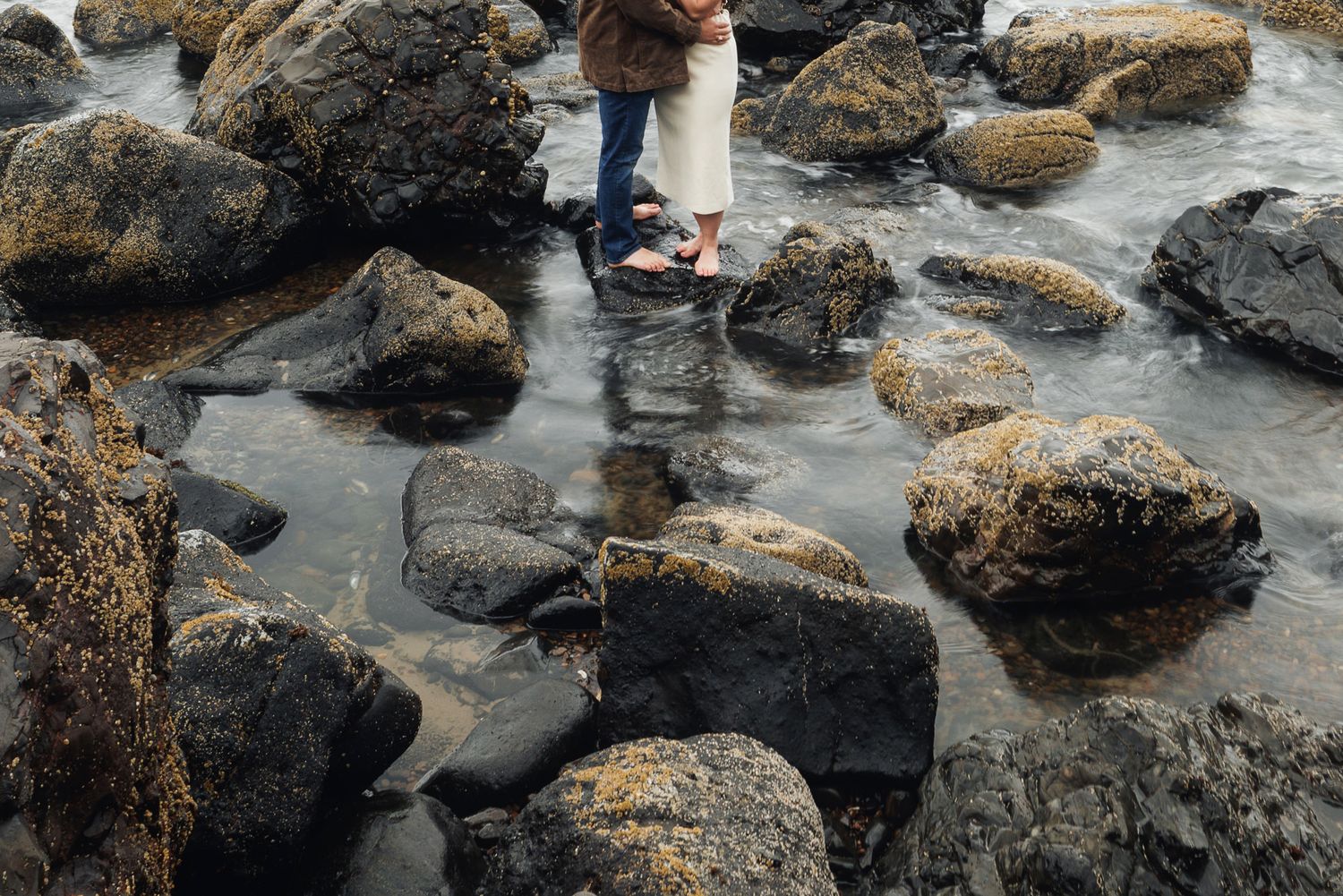 Couple standing barefoot on mossy rocks by the ocean shore, romantic coastal engagement photo.