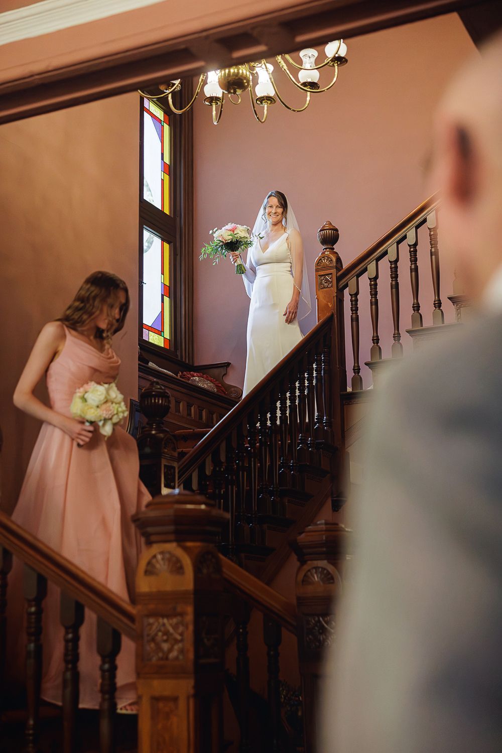 A wedding party gathers on an elegant wooden staircase with ornate railings and stained glass windows in warm lighting.