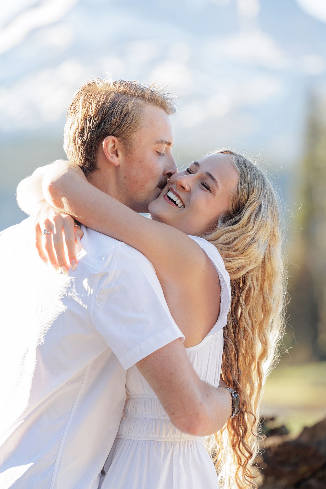 Couple in white embracing and laughing outdoors with snow-capped mountains in the background.