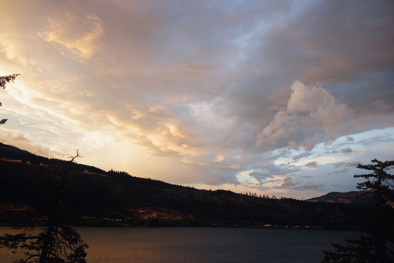 Dramatic sunset sky over a tranquil lake with silhouetted hills and trees in the foreground.