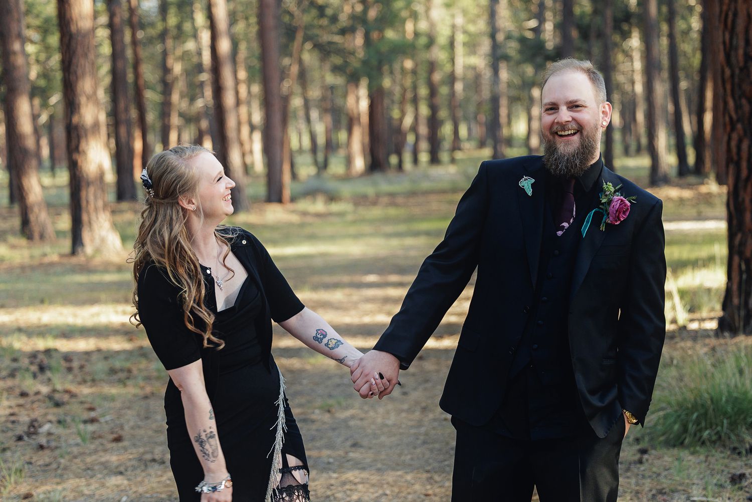 Couple in black wedding attire holding hands and smiling in a pine forest setting.
