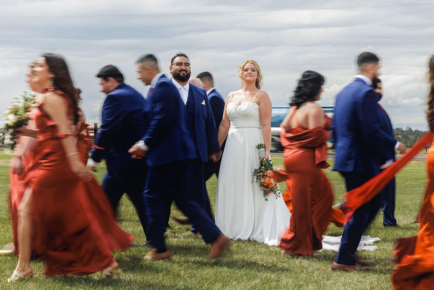 Bride and groom smile surrounded by wedding party in navy suits and rust bridesmaid dresses on green lawn.