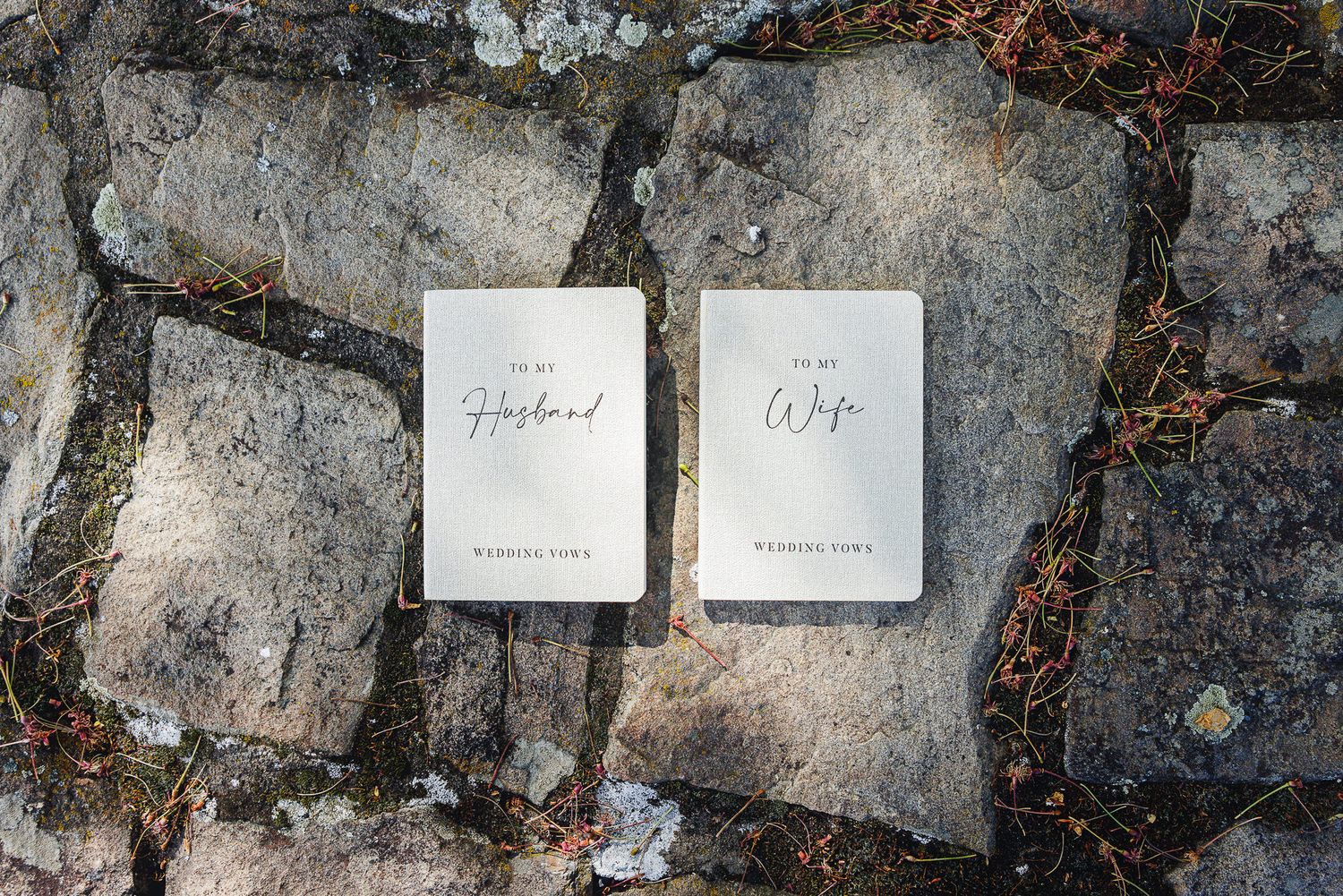 Two white wedding vow booklets labeled To My Husband and To My Wife resting on rustic stone pavers.