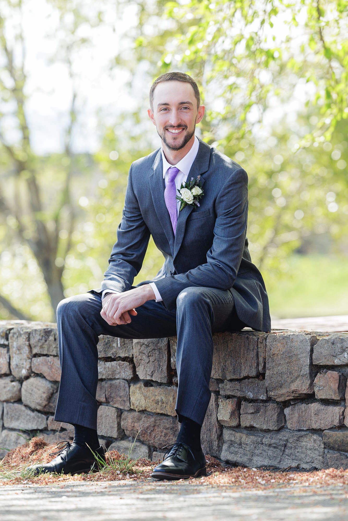 Smiling groom in navy suit with purple tie and boutonniere sitting on stone wall outdoors.