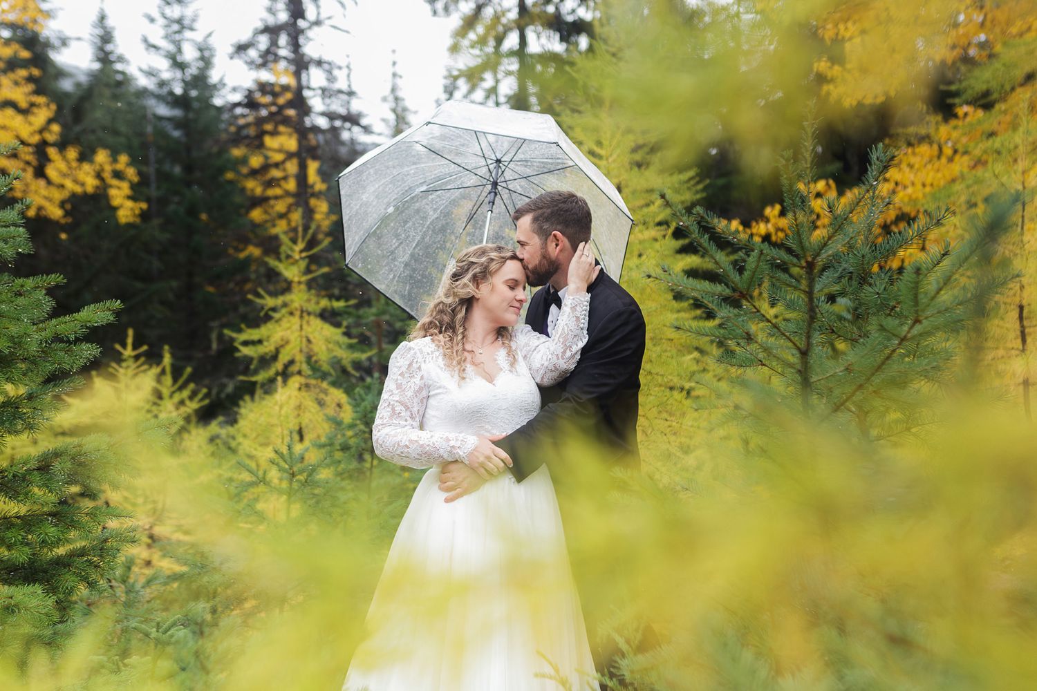 Couple in wedding attire embraces in a lush forest setting with golden autumn foliage in the background.