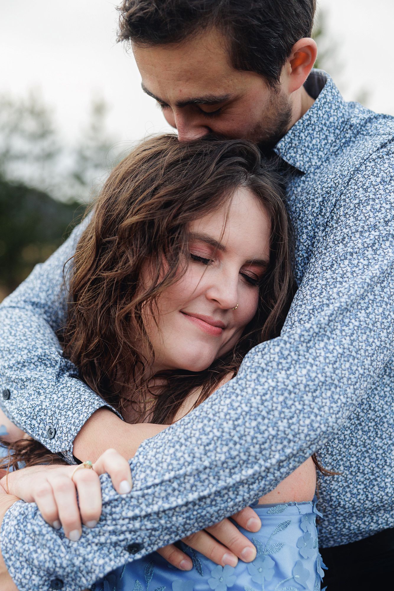 A couple embraces tenderly in a series of intimate outdoor photos, with the woman resting against his blue floral shirt.