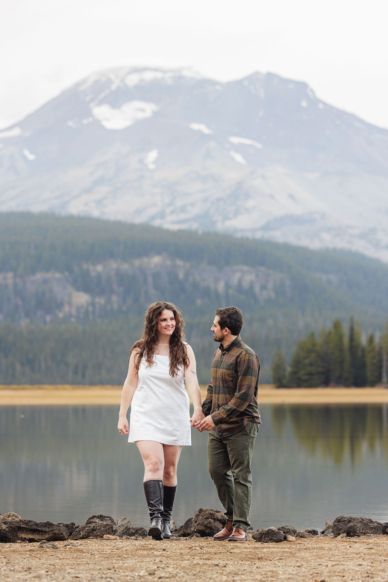 A couple stands together near a scenic mountain lake surrounded by snow-capped peaks and evergreen forests.