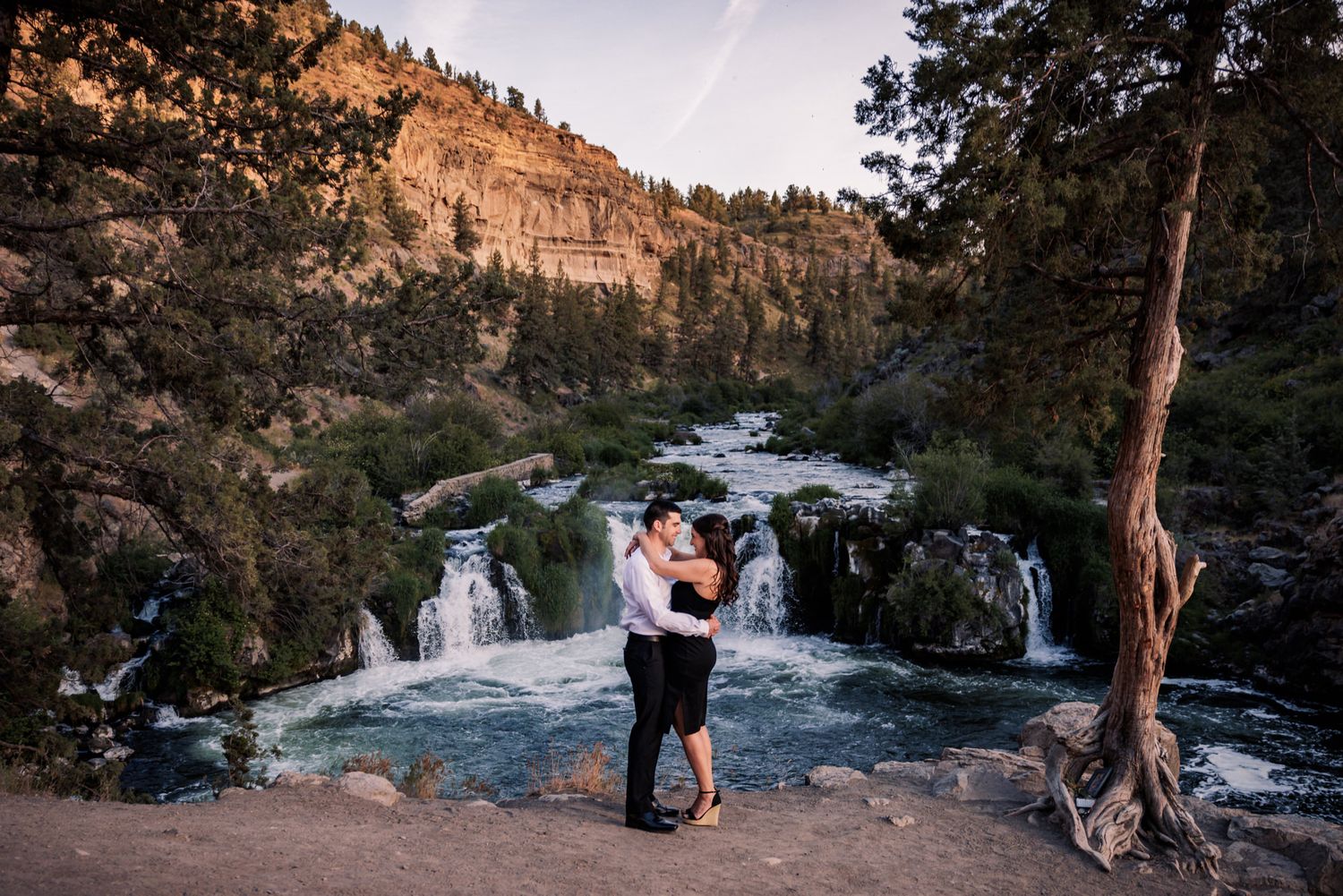 Scenic mountain engagement photo session at sunset with cascading waterfalls and rocky cliffs in the background.