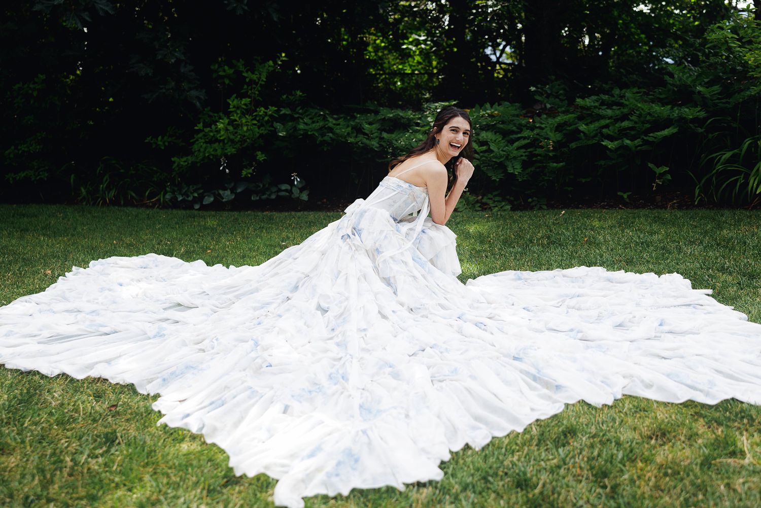 Bride in a stunning white ball gown with floral details and long train posed on green grass outdoors.