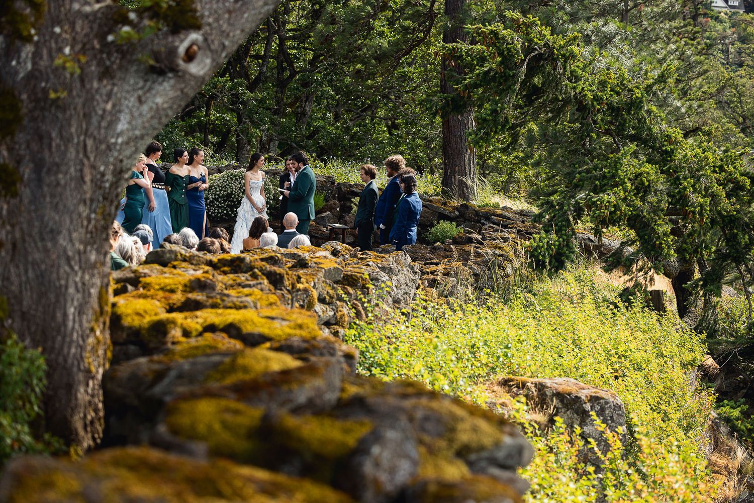 Wedding party gathered on a moss-covered stone wall surrounded by lush green trees during an outdoor ceremony.