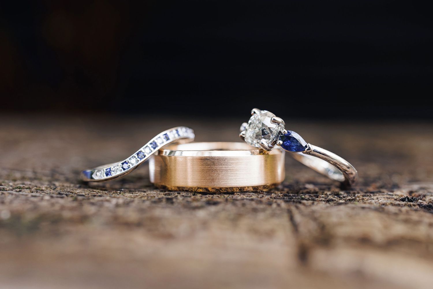 Three wedding rings on rustic wood surface, featuring gold band, diamond engagement ring, and sapphire accent ring.