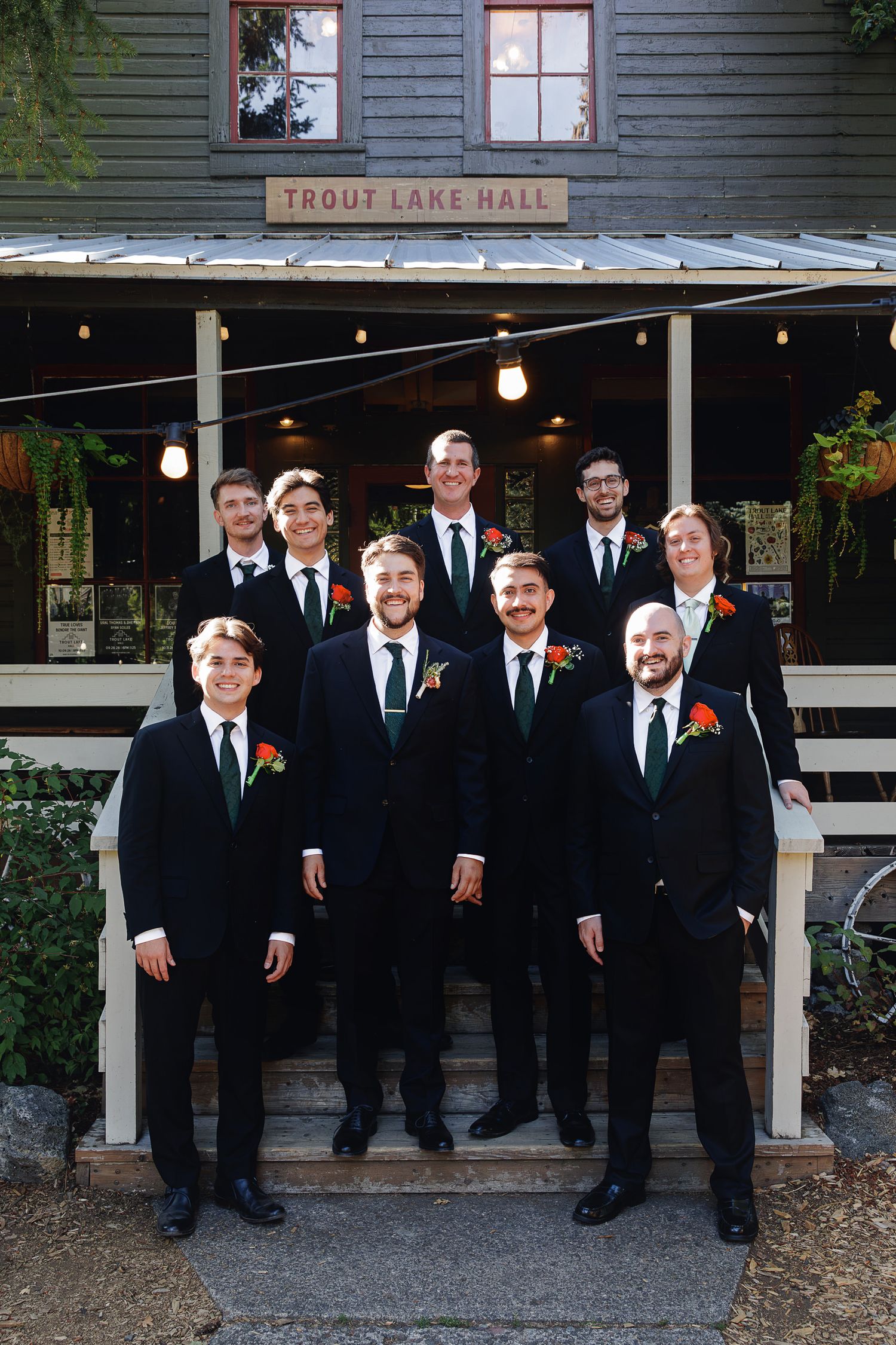 Groomsmen in black suits with red boutonnieres pose on steps outside Trout Lake Hall at a wedding.