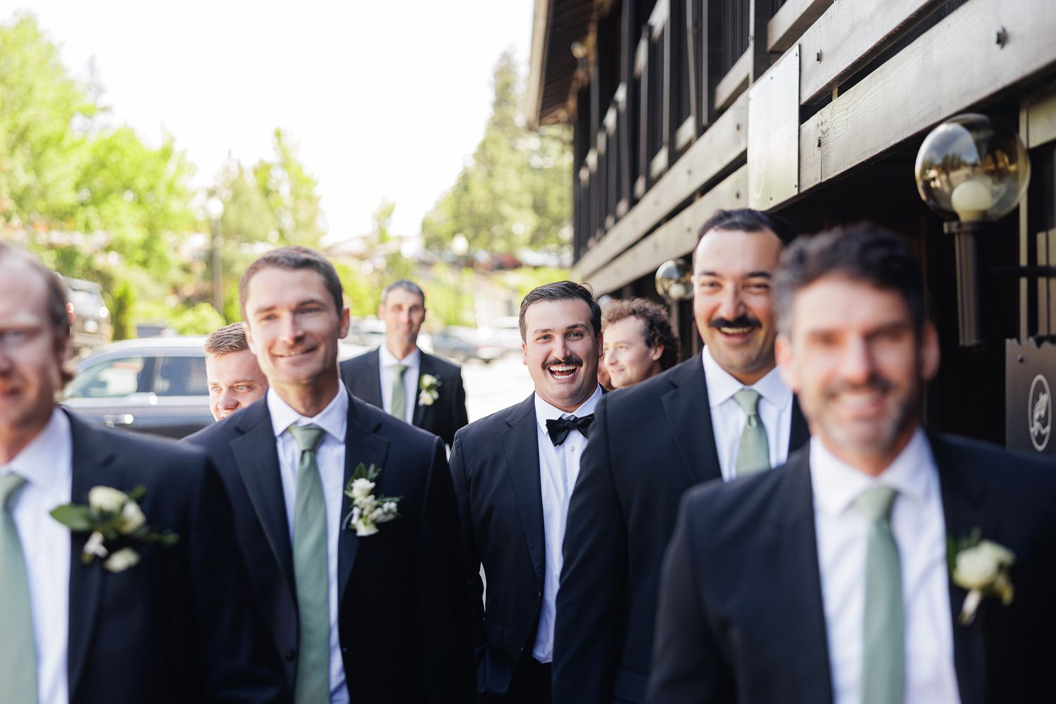 Groomsmen in dark suits with green ties and boutonnieres smiling outdoors at a wedding celebration.