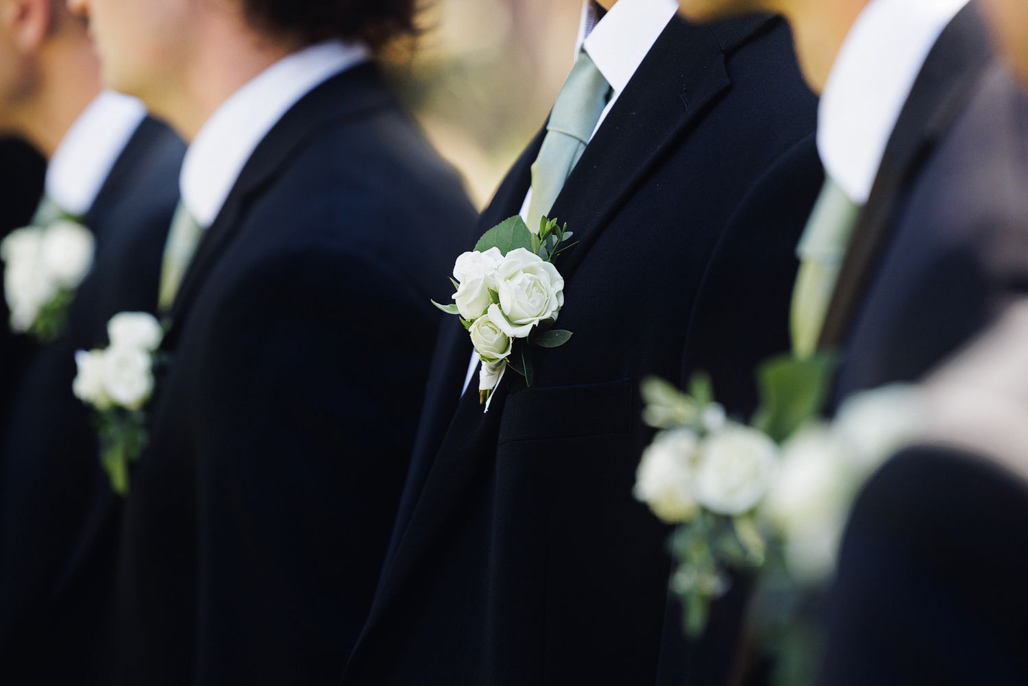 Groomsmen in black suits with white rose boutonnieres lined up at a wedding ceremony.