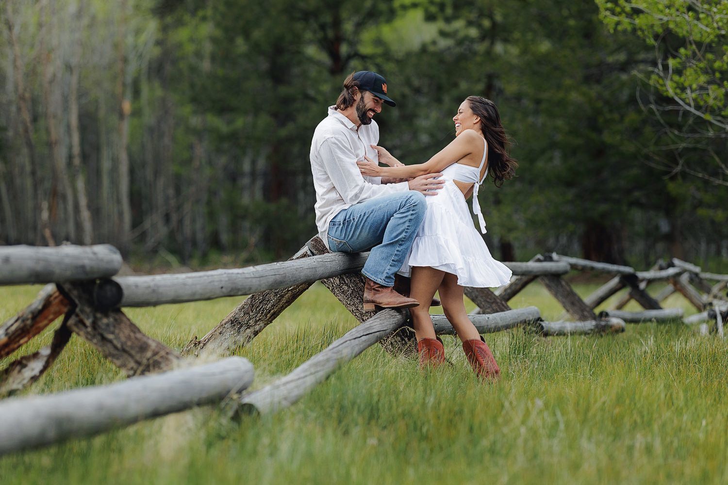 A couple sits together on a rustic wooden fence surrounded by green grass and trees in a romantic countryside setting.